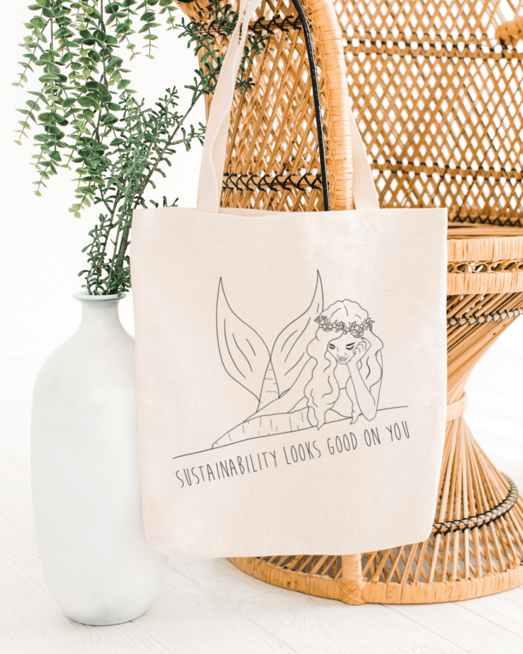 Tide + Seek sustainable swimwear tote bag hanging on a wicker bohemian style chair and pot planet. The bag is a cream colour with a mermaid on the front and slogan saying "sustainability looks good on you" across it.  